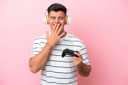 Foto de Joven hombre guapo jugando con un controlador de videojuego aislado sobre fondo rosa feliz y sonriente boca cubierta con la mano - Imagen libre de derechos