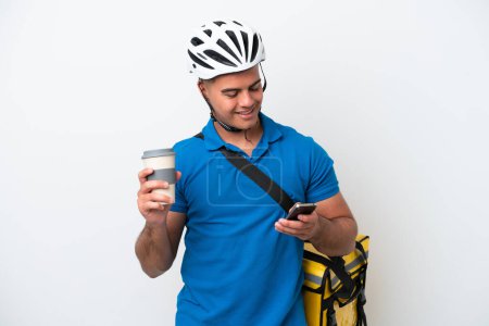 Téléchargez les photos : Jeune homme caucasien avec sac à dos thermique isolé sur fond blanc tenant du café à emporter et un mobile - en image libre de droit