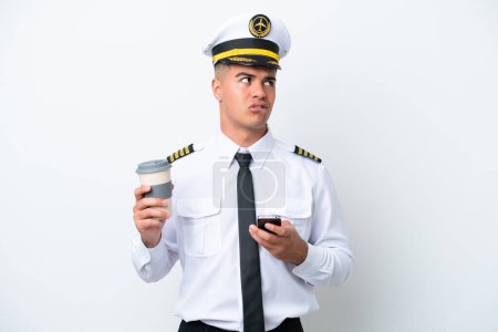Airplane pilot caucasian man isolated on white background holding coffee to take away and a mobile while thinking something