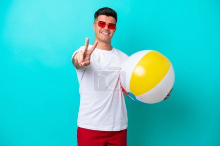 Téléchargez les photos : Jeune homme caucasien tenant une balle de plage isolé sur fond bleu souriant et montrant signe de victoire - en image libre de droit