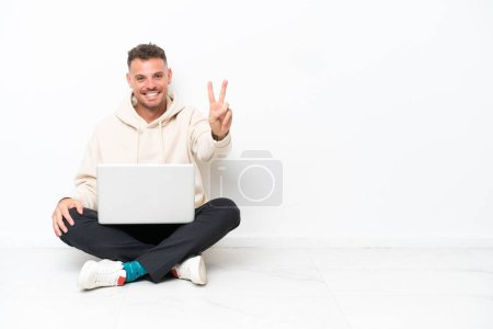 Téléchargez les photos : Jeune homme caucasien avec un ordinateur portable assis sur le sol isolé sur fond blanc souriant et montrant signe de victoire - en image libre de droit