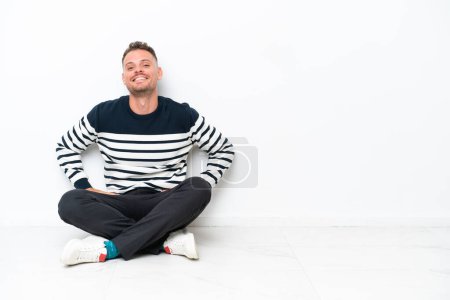 Foto de Joven sentado en el suelo aislado sobre fondo blanco posando con los brazos en la cadera y sonriendo - Imagen libre de derechos