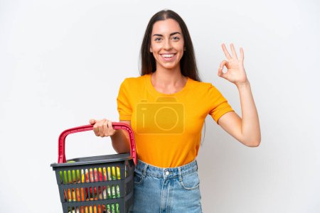 Foto de Young caucasian woman holding a shopping basket full of food isolated on white background showing ok sign with fingers - Imagen libre de derechos