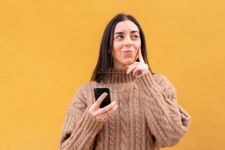 Téléchargez les photos : Jeune femme brune à l'extérieur en utilisant un téléphone mobile et en pensant - en image libre de droit