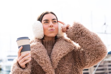 Photo for Young woman wearing winter muffs  and holding take away coffee at outdoors having doubts and with confuse face expression - Royalty Free Image