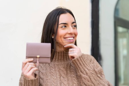 Téléchargez les photos : Jeune femme tenant un portefeuille à l'extérieur pensant une idée et regardant côté - en image libre de droit