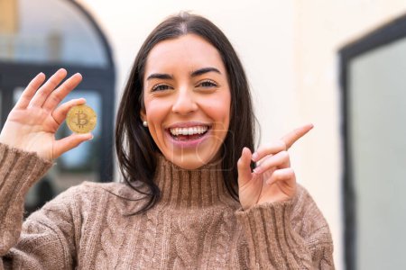 Foto de Mujer joven sosteniendo un Bitcoin al aire libre sorprendido y señalando el dedo hacia un lado - Imagen libre de derechos