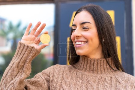 Téléchargez les photos : Jeune femme tenant un Bitcoin à l'extérieur avec une expression heureuse - en image libre de droit