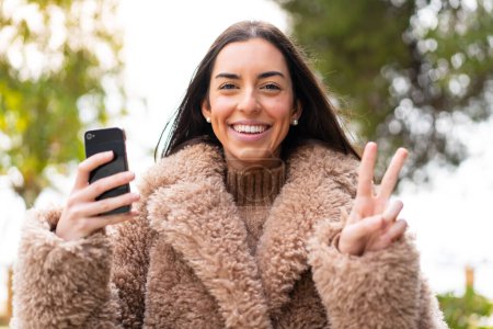 Foto de Mujer joven usando el teléfono móvil al aire libre sonriendo y mostrando la señal de victoria - Imagen libre de derechos