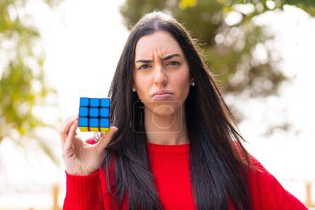 Téléchargez les photos : Jeune femme tenant un cube de puzzle tridimensionnel à l'extérieur avec une expression triste - en image libre de droit