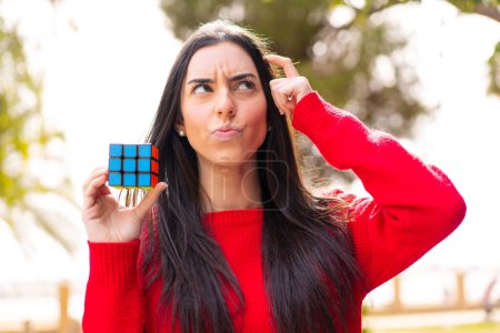 Photo for Young woman holding a three dimensional puzzle cube at outdoors having doubts and with confuse face expression - Royalty Free Image