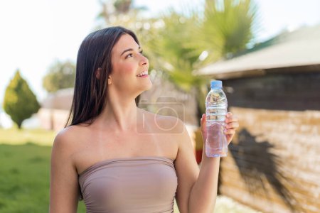 Photo for Young Brazilian woman with a bottle of water at outdoors looking up while smiling - Royalty Free Image