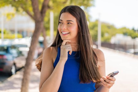 Foto de Joven mujer brasileña bonita usando el teléfono móvil al aire libre pensando en una idea y mirando al lado - Imagen libre de derechos