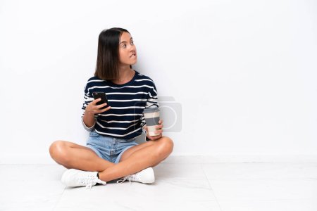 Téléchargez les photos : Jeune femme caucasienne assise sur le sol isolée sur fond blanc tenant du café à emporter et un mobile tout en pensant quelque chose - en image libre de droit