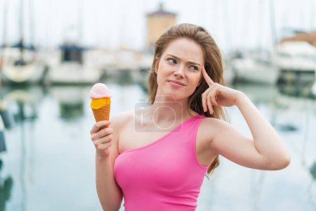 Foto de Joven pelirroja con un helado de corneta al aire libre que tiene dudas y con expresión de cara confusa - Imagen libre de derechos