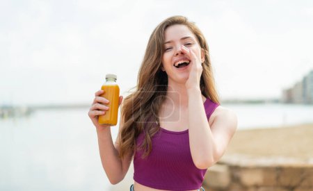 Téléchargez les photos : Jeune fille rousse tenant un jus d'orange à l'extérieur criant avec la bouche grande ouverte - en image libre de droit