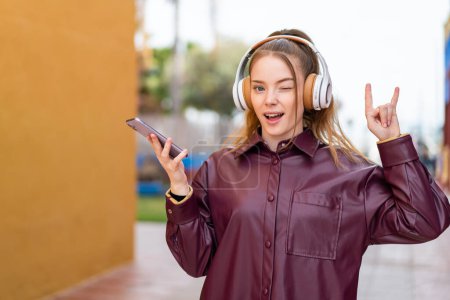 Foto de Joven chica bonita al aire libre escuchando música con un móvil haciendo un gesto de rock - Imagen libre de derechos