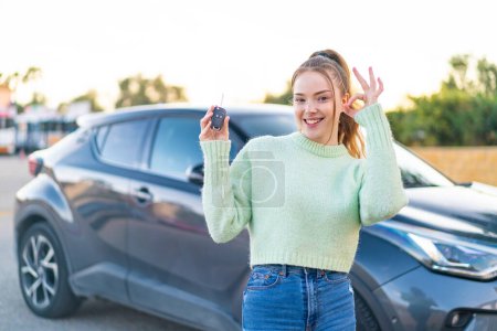 Téléchargez les photos : Jeune jolie fille tenant des clés de voiture à l'extérieur montrant ok signe avec les doigts - en image libre de droit