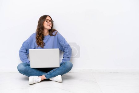 Téléchargez les photos : Jeune femme caucasienne avec un ordinateur portable assis sur le sol isolé sur fond blanc posant avec les bras à la hanche et souriant - en image libre de droit