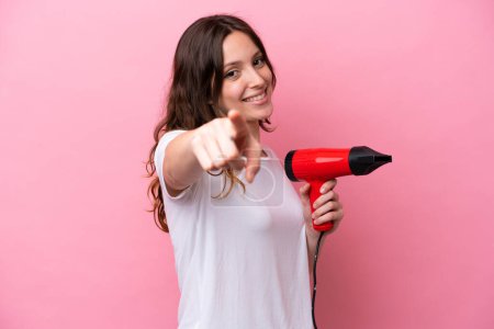 Téléchargez les photos : Jeune femme caucasienne tenant un sèche-cheveux isolé sur fond rose pointe doigt à vous avec une expression confiante - en image libre de droit