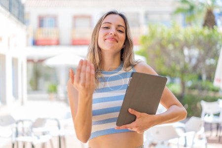 Téléchargez les photos : Jeune jolie femme tenant une tablette à l'extérieur invitant à venir avec la main. Heureux que tu sois venu - en image libre de droit