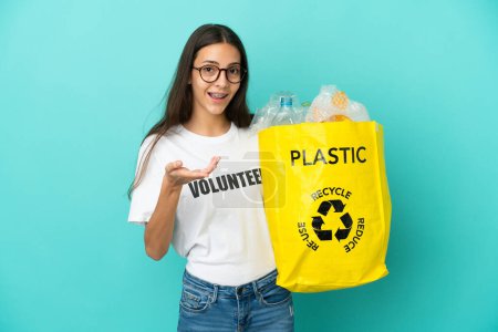 Téléchargez les photos : Jeune fille française tenant un sac rempli de bouteilles en plastique pour recycler les mains tendues sur le côté pour inviter à venir - en image libre de droit