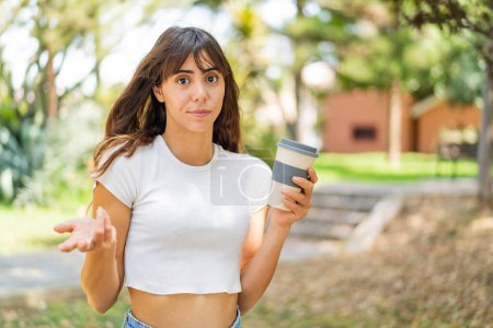 Junge Frau hält im Freien einen Kaffee zum Mitnehmen und macht beim Anheben der Schultern eine Geste des Zweifels