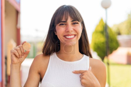 Foto de Mujer joven sosteniendo un Bitcoin al aire libre con expresión facial sorpresa - Imagen libre de derechos