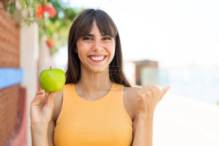 Téléchargez les photos : Jeune femme avec une pomme à l'extérieur pointant vers le côté pour présenter un produit - en image libre de droit