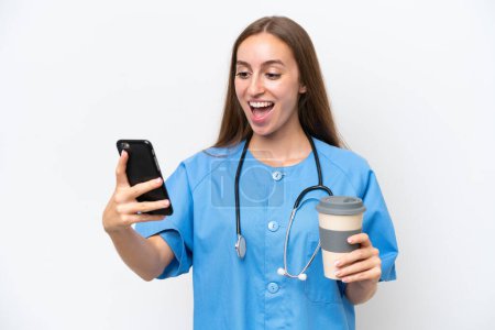 Foto de Young nurse woman over isolated on white background holding coffee to take away and a mobile - Imagen libre de derechos