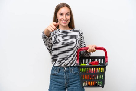 Photo for Young Rumanian woman holding a shopping basket full of food isolated on white background surprised and pointing front - Royalty Free Image