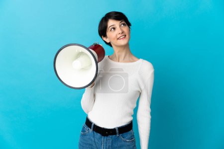 Téléchargez les photos : Femme aux cheveux courts isolés sur fond bleu tenant un mégaphone et levant les yeux tout en souriant - en image libre de droit