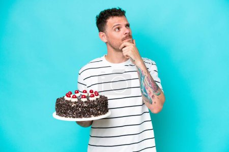 Foto de Young caucasian man holding birthday cake isolated on blue background having doubts - Imagen libre de derechos