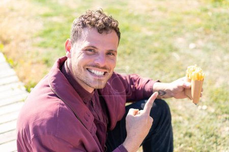Foto de Joven hombre caucásico sosteniendo papas fritas al aire libre y señalándolo - Imagen libre de derechos