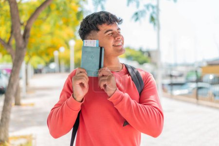 Téléchargez les photos : Jeune homme hispanique à l'extérieur tenant un passeport avec une expression heureuse - en image libre de droit
