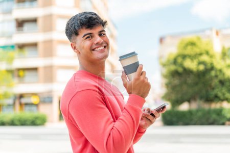 Photo for Young hispanic man at outdoors using mobile phone and holding a coffee with happy expression - Royalty Free Image