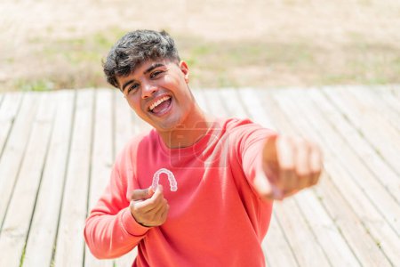 Téléchargez les photos : Jeune homme hispanique tenant des bretelles invisibles à l'extérieur pointe du doigt vers vous avec une expression confiante - en image libre de droit