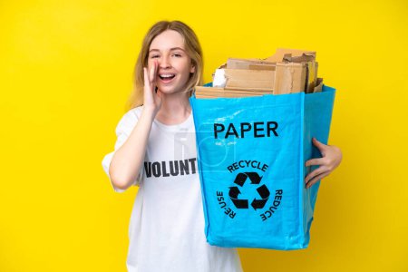 Blonde English young girl holding a recycling bag full of paper to recycle isolated on yellow background shouting with mouth wide open