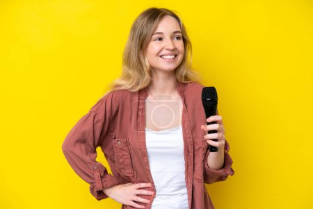 Foto de Joven cantante caucásica recogiendo un micrófono aislado sobre fondo amarillo posando con los brazos en la cadera y sonriendo - Imagen libre de derechos