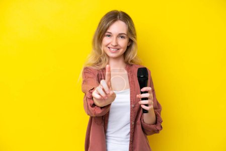Téléchargez les photos : Jeune chanteuse caucasienne ramasser un microphone isolé sur fond jaune montrant et soulevant un doigt - en image libre de droit