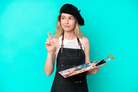 Photo for Young artist caucasian woman holding a palette isolated on blue background with fingers crossing and wishing the best - Royalty Free Image