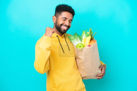 Photo for Young Brazilian man taking a bag of takeaway food isolated on blue background celebrating a victory - Royalty Free Image