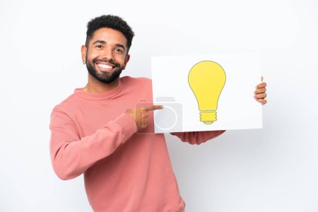 Photo for Young Brazilian man isolated on white background holding a placard with bulb icon and pointing it - Royalty Free Image
