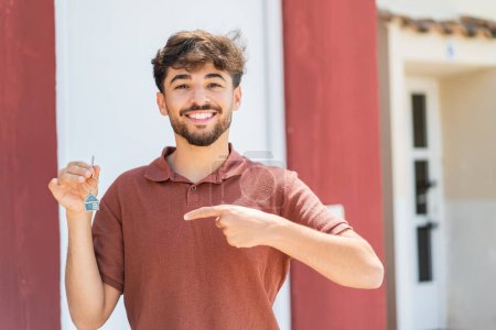 Téléchargez les photos : Jeune homme beau arabe tenant les clés de la maison à l'extérieur et le pointant - en image libre de droit