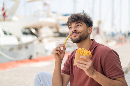Foto de Joven árabe guapo al aire libre tomando papas fritas y pensando - Imagen libre de derechos