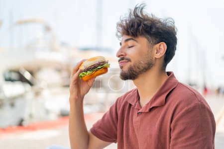 Foto de Joven árabe guapo al aire libre sosteniendo una hamburguesa - Imagen libre de derechos
