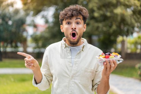Téléchargez les photos : Jeune homme beau arabe tenant un bol de fruits à l'extérieur surpris et pointant côté - en image libre de droit
