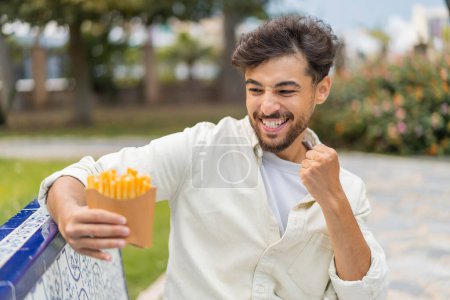 Foto de Joven árabe guapo sosteniendo patatas fritas al aire libre celebrando una victoria - Imagen libre de derechos