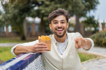 Photo for Young Arabian handsome man holding fried chips at outdoors points finger at you with a confident expression - Royalty Free Image
