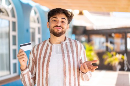 Handsome Arab man holding a credit card at outdoors making doubts gesture while lifting the shoulders